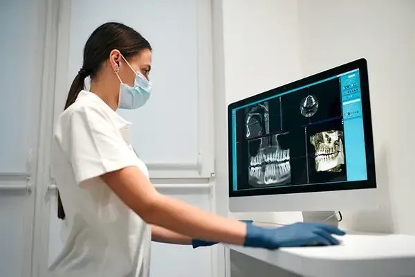 Dental professional in a mask and gloves examining high-resolution dental images on a computer for full-mouth restoration planning.