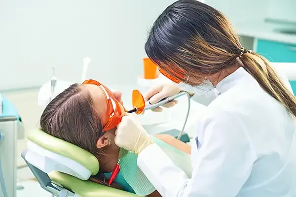 A dentist applying a dental sealant to a young patient's teeth using a curing light while the patient wears protective glasses in a dental clinic.