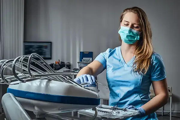 A dental assistant in a blue uniform and face mask organizing surgical dental tools in a clinic.