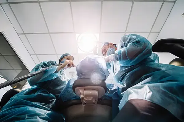 A low-angle view of dental surgeons in protective clothing working on a patient under bright surgical lights during a dental procedure.